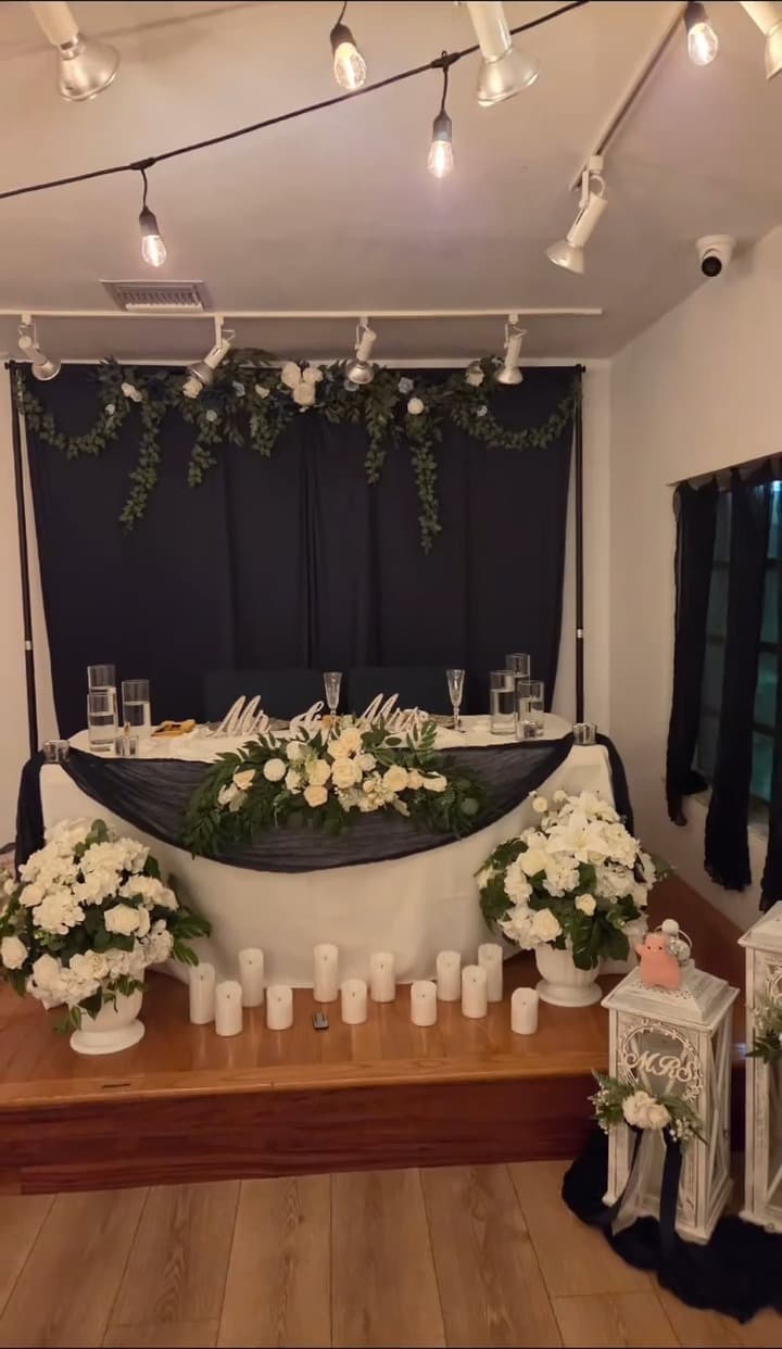 Sweetheart table with black draped backdrop, white floral garland and rows of pillar candles